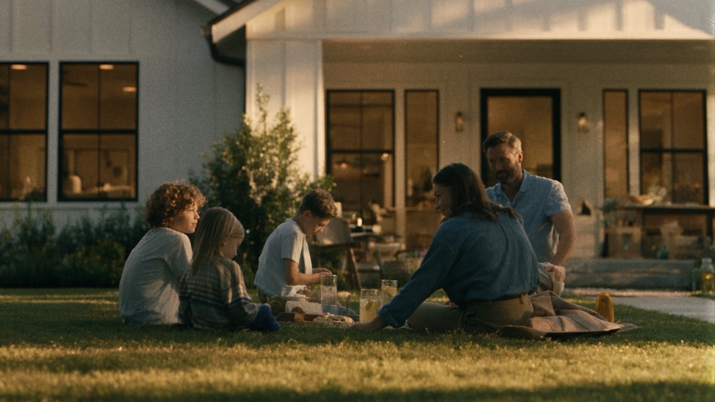 A family sitting on the lawn outside a lit house at golden hour